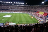 A large soccer stadium filled with a crowd of spectators during a nighttime event. The field is well-lit, and there are teams or players in the center circle preparing for a match. A section of fans is displaying a large red and yellow banner, and the atmosphere is lively with numerous people visible in the stands.