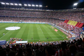 A large soccer stadium filled with a crowd of spectators during a nighttime event. The field is well-lit, and there are teams or players in the center circle preparing for a match. A section of fans is displaying a large red and yellow banner, and the atmosphere is lively with numerous people visible in the stands.