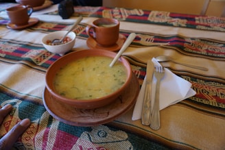 A table is set with a colorful woven tablecloth featuring traditional patterns. On the table, there is a brown ceramic bowl containing a thick, greenish-yellow soup with herbs, and a wooden spoon inside it. Nearby, there is a small white bowl with a spoon, containing a mix of chopped vegetables or condiments, and a brown ceramic mug on a saucer. A set of silverware, including a knife and fork, is placed on a white napkin.