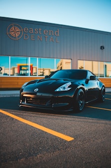 A sleek black sports car is parked in front of a commercial building with a sign for 'east end DENTAL.' The car has a glossy finish and is positioned on an asphalt parking lot, marked with yellow lines. The building has large windows reflecting the surrounding area.