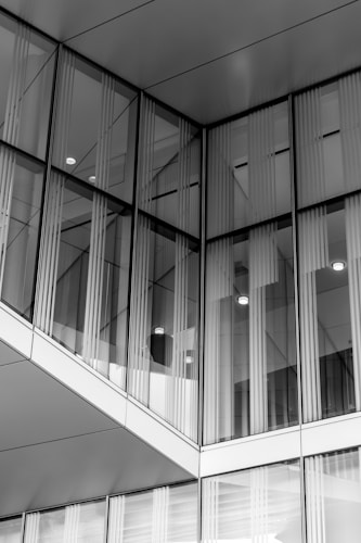 A modern building facade featuring large glass panels reflecting the surroundings. The glass elements are framed with sleek metal lines, and there are vertical blinds visible through the glass. The structure has a geometric, angular design.