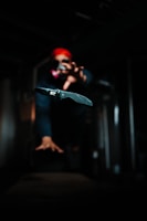 A folding knife is prominently displayed in sharp focus with a blurred background. A person wearing a red headscarf and sunglasses appears to be reaching towards the knife, adding a sense of action or drama. The setting is dimly lit, creating a mysterious and tense atmosphere.