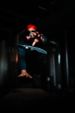 A folding knife is prominently displayed in sharp focus with a blurred background. A person wearing a red headscarf and sunglasses appears to be reaching towards the knife, adding a sense of action or drama. The setting is dimly lit, creating a mysterious and tense atmosphere.