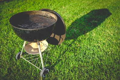 A charcoal grill lies on a well-manicured lawn, casting a long shadow in the sunlight. The grill is black with a gridiron surface and has a circular design with metal legs and wheels for support.