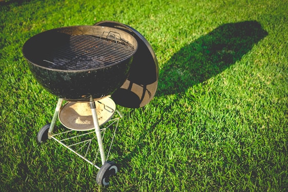 A charcoal grill lies on a well-manicured lawn, casting a long shadow in the sunlight. The grill is black with a gridiron surface and has a circular design with metal legs and wheels for support.