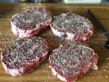 Four raw, marbled steak cuts are seasoned with salt and pepper, arranged on a wooden cutting board alongside a black-handled knife.