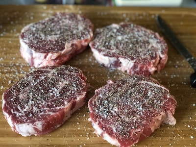 Four raw, marbled steak cuts are seasoned with salt and pepper, arranged on a wooden cutting board alongside a black-handled knife.