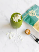 A green smoothie topped with coconut flakes and seeds is placed on a white marble surface. Nearby, a teal bag with a botanical design and a measuring spoon with a brown powder are visible.