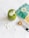 A green smoothie topped with coconut flakes and seeds is placed on a white marble surface. Nearby, a teal bag with a botanical design and a measuring spoon with a brown powder are visible.