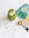 A green smoothie topped with coconut flakes and seeds is placed on a white marble surface. Nearby, a teal bag with a botanical design and a measuring spoon with a brown powder are visible.