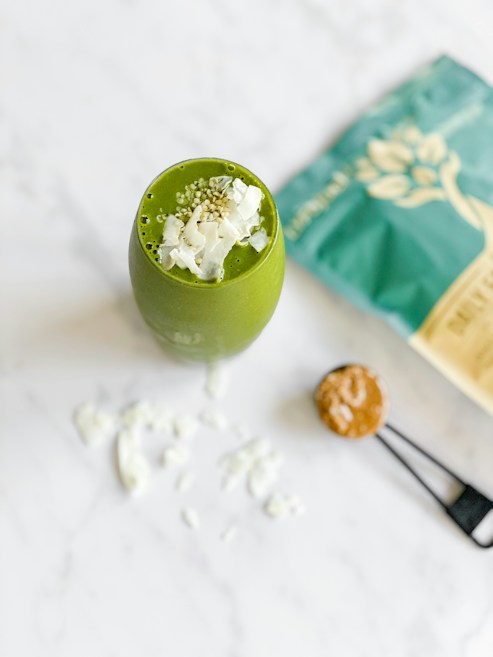 A green smoothie topped with coconut flakes and seeds is placed on a white marble surface. Nearby, a teal bag with a botanical design and a measuring spoon with a brown powder are visible.