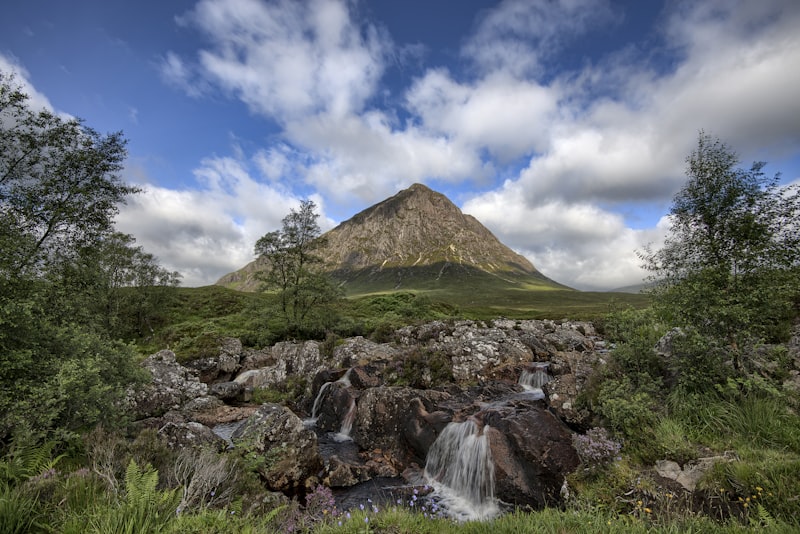 Pyramid-shaped rocky mountain rising above green moorland with a small waterfall tumbling over boulders in the foreground under a blue sky with clouds
