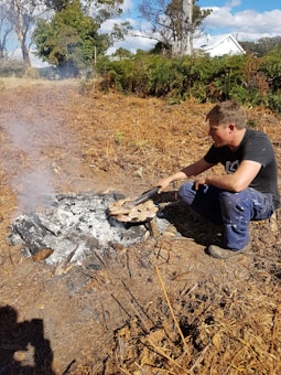 A person crouches beside a small campfire, tending to food cooking on a metal grill placed over ash-covered logs. Surrounding the area are dry brown ferns and trees, with a glimpse of a house in the background under a partly cloudy sky.