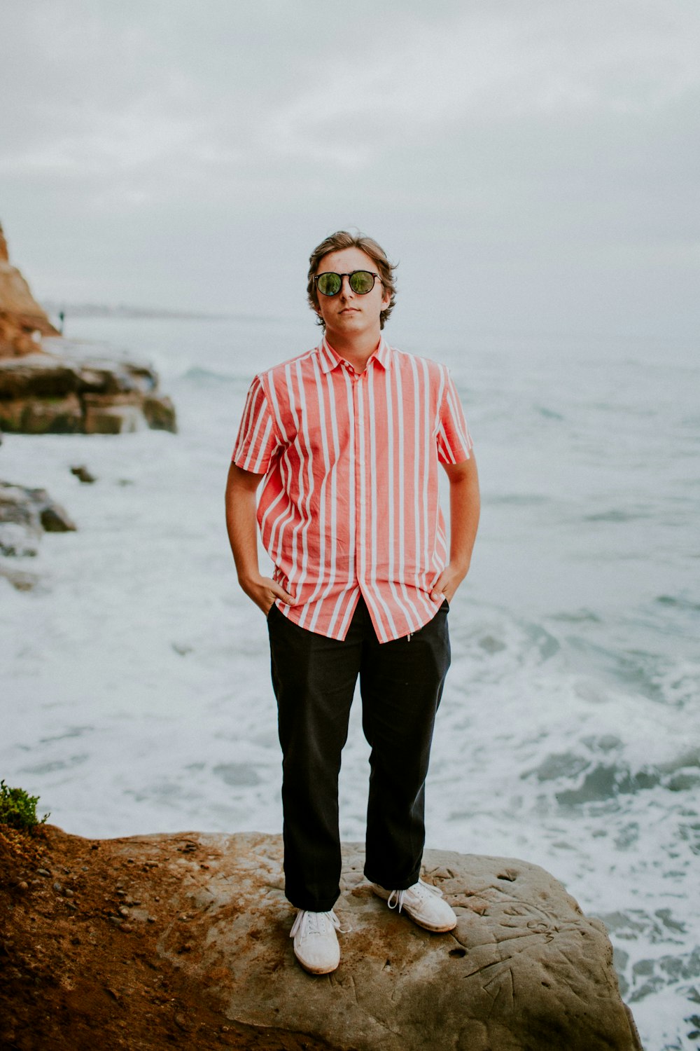 man in pink and white striped button up shirt standing on rock formation near sea during