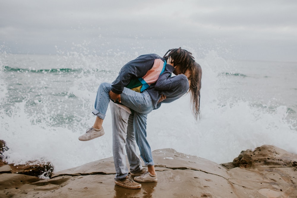 woman in blue jacket and gray pants carrying woman in blue jacket on beach during daytime