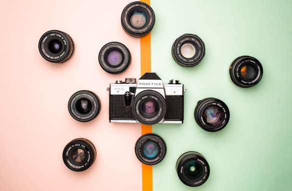A vintage camera is surrounded by multiple camera lenses arranged in a circular pattern. The background is split into two pastel colors, pink on the left and green on the right, divided by a thin orange line.