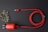 A red braided charging cable is neatly coiled next to a charger with a plug, set against a dark background. On the left, a sprig of dried white flowers adds an organic touch.