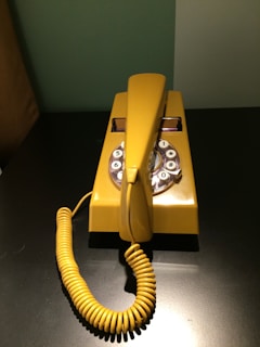 A vintage rotary telephone in bright yellow sits on a dark surface. The phone features a coiled cord and a dial with visible numbers and symbols. The background is a two-tone wall in muted green and grey.