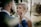 A bride and groom share a tender moment in a lush, green outdoor setting. The groom gently holds the bride's face, both gazing lovingly into each other's eyes. The background is softly blurred with trees providing a serene atmosphere.