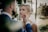A bride and groom share a tender moment in a lush, green outdoor setting. The groom gently holds the bride's face, both gazing lovingly into each other's eyes. The background is softly blurred with trees providing a serene atmosphere.
