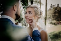 A bride and groom share a tender moment in a lush, green outdoor setting. The groom gently holds the bride's face, both gazing lovingly into each other's eyes. The background is softly blurred with trees providing a serene atmosphere.