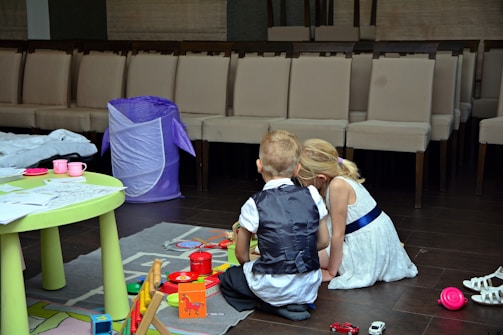 Two children, a boy and a girl, are sitting on the floor playing with toys. The setting appears to be indoors, with rows of chairs in the background suggesting a waiting area or event space. A table with papers and toy cups, as well as a purple hamper, are nearby.