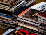 A collection of stacked compact disc cases, some with visible album artwork including text and images. The cases vary in color and design, creating a diverse and visually engaging scene.