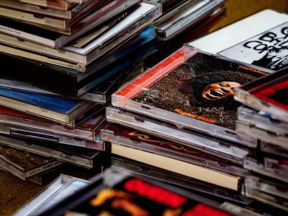 A collection of stacked compact disc cases, some with visible album artwork including text and images. The cases vary in color and design, creating a diverse and visually engaging scene.