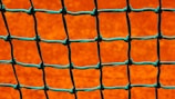 A close-up view of a green net with square openings set against a bright orange background. The net is neatly tied with visible knots, creating a grid-like pattern.