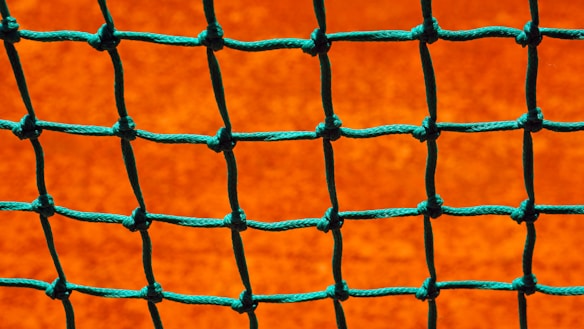 A close-up view of a green net with square openings set against a bright orange background. The net is neatly tied with visible knots, creating a grid-like pattern.