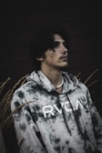 A person is wearing a tie-dye hoodie with the letters RVCA printed on it. The person is looking upwards, and the setting includes a dark background with tall grass or wheat surrounding them.