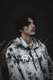 A person is wearing a tie-dye hoodie with the letters RVCA printed on it. The person is looking upwards, and the setting includes a dark background with tall grass or wheat surrounding them.