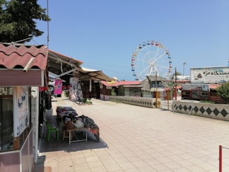 An outdoor market area features stalls offering various goods including clothing items displayed on tables and hanging. In the background, a large Ferris wheel is prominent against a clear blue sky. The area is paved with stone tiles, and there are several structures with red roofs. Sparse greenery is visible around the periphery.