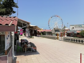 An outdoor market area features stalls offering various goods including clothing items displayed on tables and hanging. In the background, a large Ferris wheel is prominent against a clear blue sky. The area is paved with stone tiles, and there are several structures with red roofs. Sparse greenery is visible around the periphery.