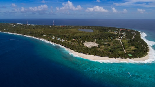 An aerial view of a lush, green island with dense vegetation surrounded by clear turquoise waters and white sandy beaches. The coastline curves gently with waves crashing against it, and there are several structures visible, including buildings and a road running parallel to the shore. The sky is mostly clear with a few scattered clouds.