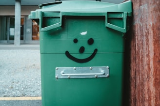 A green garbage bin with a smiley face graffiti on it is positioned near a wooden surface outdoors. The background features a gray gravel ground and a building entrance.