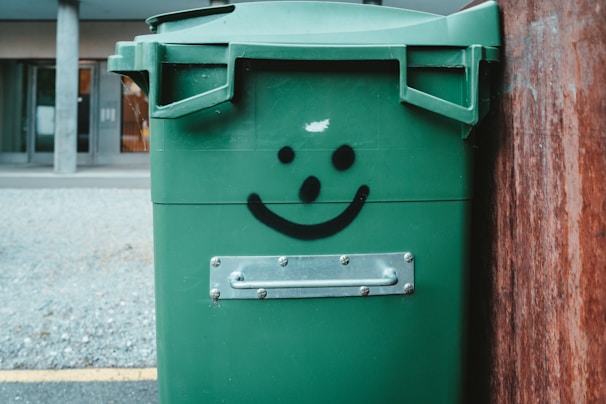 A green garbage bin with a smiley face graffiti on it is positioned near a wooden surface outdoors. The background features a gray gravel ground and a building entrance.