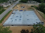 Aerial view of a construction site with a partially built structure, surrounded by dirt and machinery. The building framework is visible, and several steel beams are laid out on the concrete foundation. Roads and a few buildings are seen in the background.