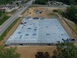Aerial view of a construction site with a partially built structure, surrounded by dirt and machinery. The building framework is visible, and several steel beams are laid out on the concrete foundation. Roads and a few buildings are seen in the background.