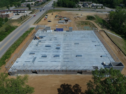 Aerial view of a construction site with a partially built structure, surrounded by dirt and machinery. The building framework is visible, and several steel beams are laid out on the concrete foundation. Roads and a few buildings are seen in the background.