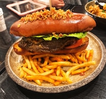 A large sandwich with a brown, toasted bun topped with crumbs, lettuce, tomato, and what seems to be a beef patty, placed on a ceramic plate with a side of seasoned French fries. In the background, a person is visible along with a tablet device and a bowl of fried food.