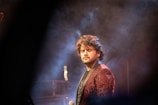 A person with closed eyes stands in a dimly lit environment surrounded by smoke or mist. The individual is holding a microphone and is wearing a dark patterned jacket. The background is blurred, creating a dreamy atmosphere.