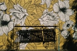 A wall mural features intricate black and white floral illustrations against a yellow background. In front of the mural, there is a metal structure casting shadows, which adds a layer of complexity to the artwork.