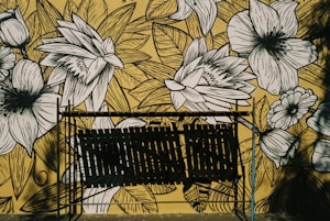 A wall mural features intricate black and white floral illustrations against a yellow background. In front of the mural, there is a metal structure casting shadows, which adds a layer of complexity to the artwork.