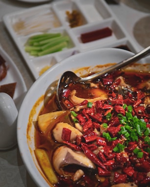 A bowl filled with a spicy dish featuring fish, garnished with a generous amount of chopped red chili peppers and green onions. The dish is accompanied by side servings of various raw vegetables such as celery sticks and what appears to be strips of tofu or noodle. A silver ladle is partially submerged in the rich, red broth.