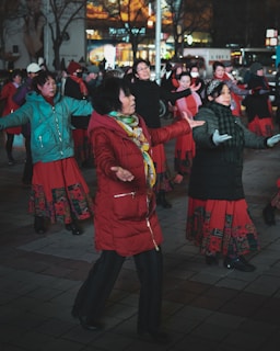 A group of people are participating in an outdoor group dance session, wearing colorful and warm clothing appropriate for a cool evening. The setting appears lively and social, with everyone seeming to enjoy themselves, suggesting a community or recreational event.
