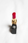 A red lipstick with a gold and black tube, featuring a recognizable logo on the base, set against a soft white background.