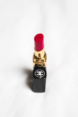 A red lipstick with a gold and black tube, featuring a recognizable logo on the base, set against a soft white background.