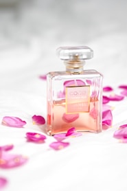 A clear glass perfume bottle with a light pink liquid inside is surrounded by scattered pink rose petals on a soft white surface. The bottle has a square shape with a gold label and a transparent top, conveying a sense of luxury and elegance.