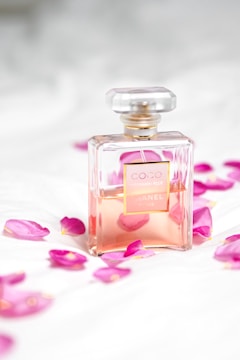 A clear glass perfume bottle with a light pink liquid inside is surrounded by scattered pink rose petals on a soft white surface. The bottle has a square shape with a gold label and a transparent top, conveying a sense of luxury and elegance.